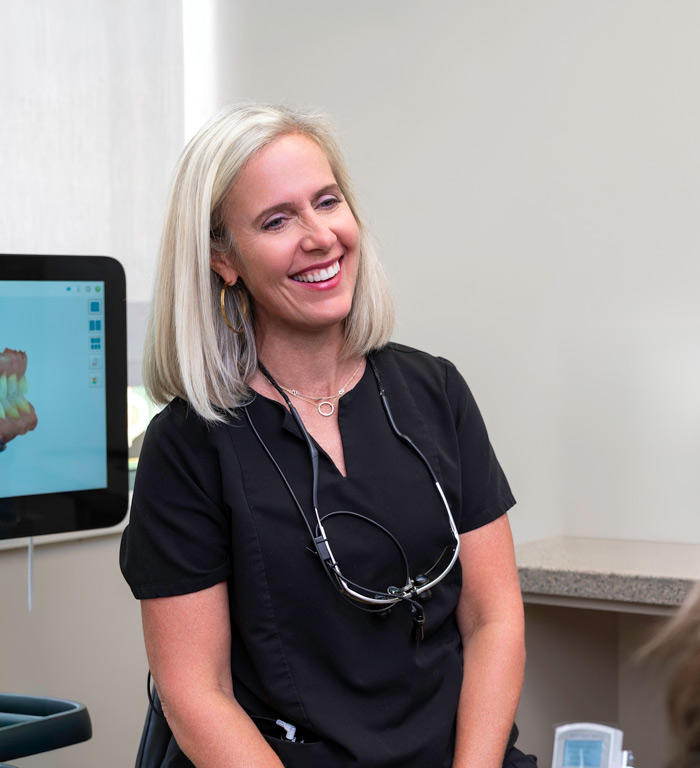 McKnight Signature Dental Smiling dental professional in black scrubs with loupes around her neck, seated in a dental treatment room.