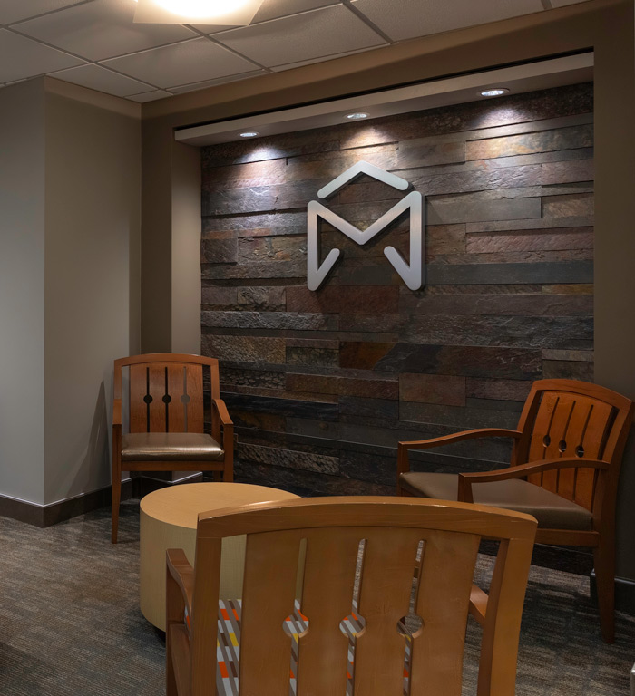 McKnight Signature Dental Dental office waiting area with wooden chairs, a small table, and a geometric 'M' logo mounted on a stone wall.