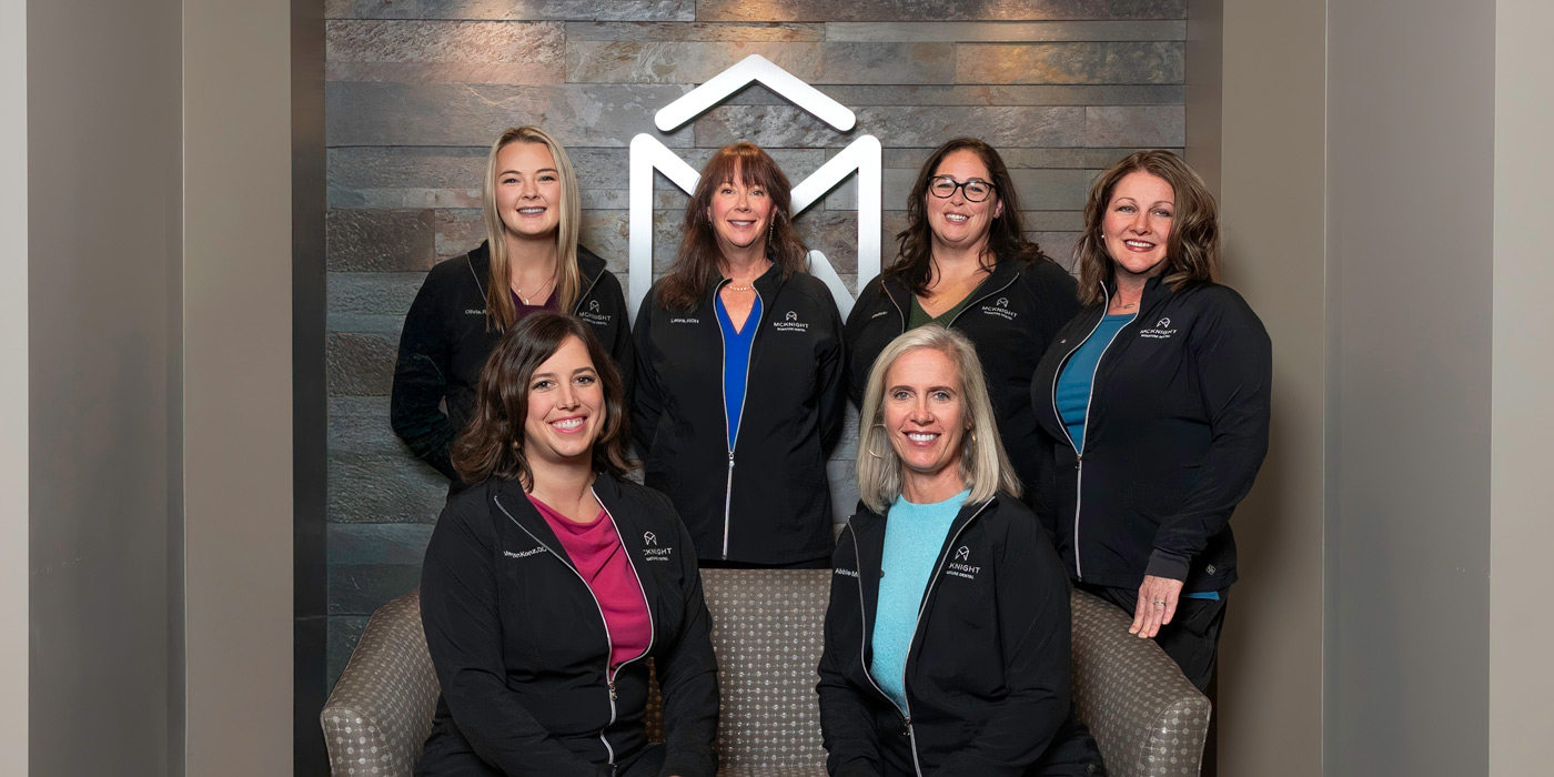 McKnight Signature Dental Group photo of seven dental team members in matching black jackets with the McKnight Signature Dental logo, posing in front of a stone wall with a large geometric 'M' logo.