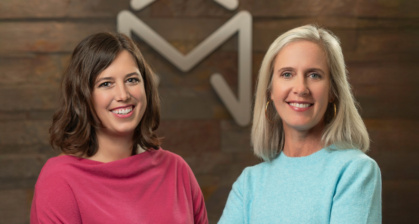 McKnight Signature Dental Two smiling women standing in front of a wooden wall with a large geometric 'M' logo in the background.
